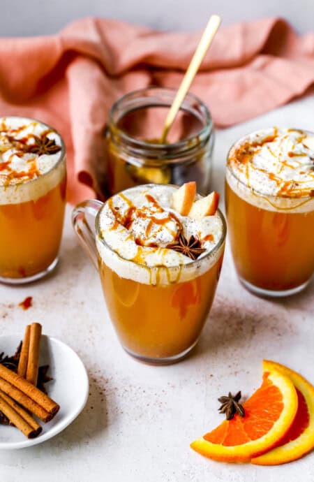 Three mugs of spiced cider with whipped cream and caramel sauce surrounded by a coral napkin