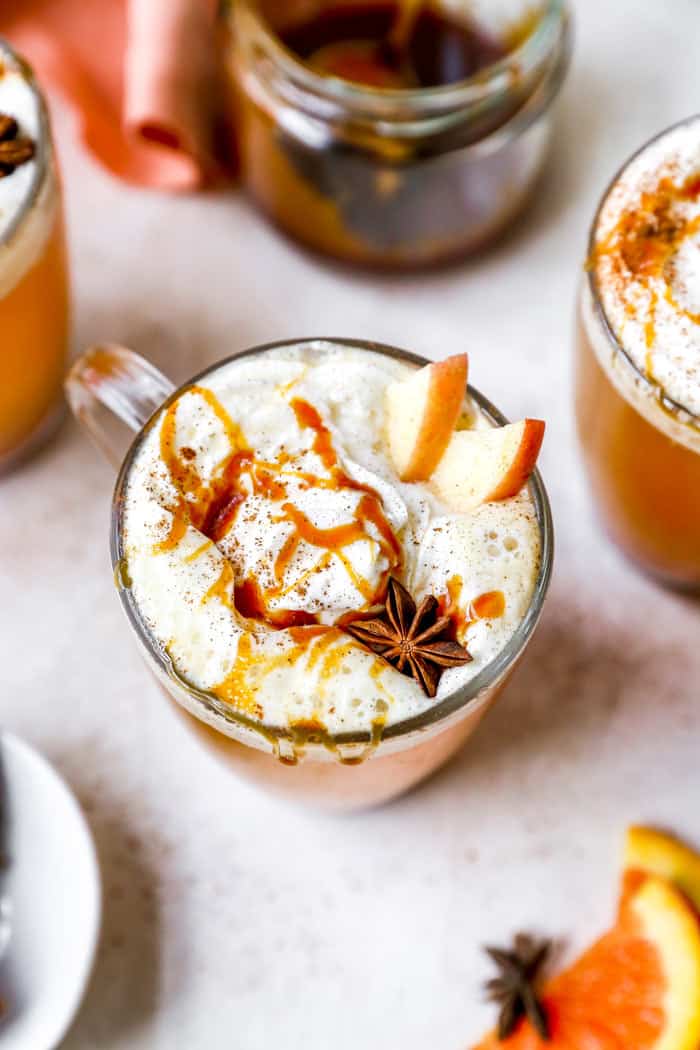 Caramel sauce in the background with three spiced apple cider mugs ready to serve