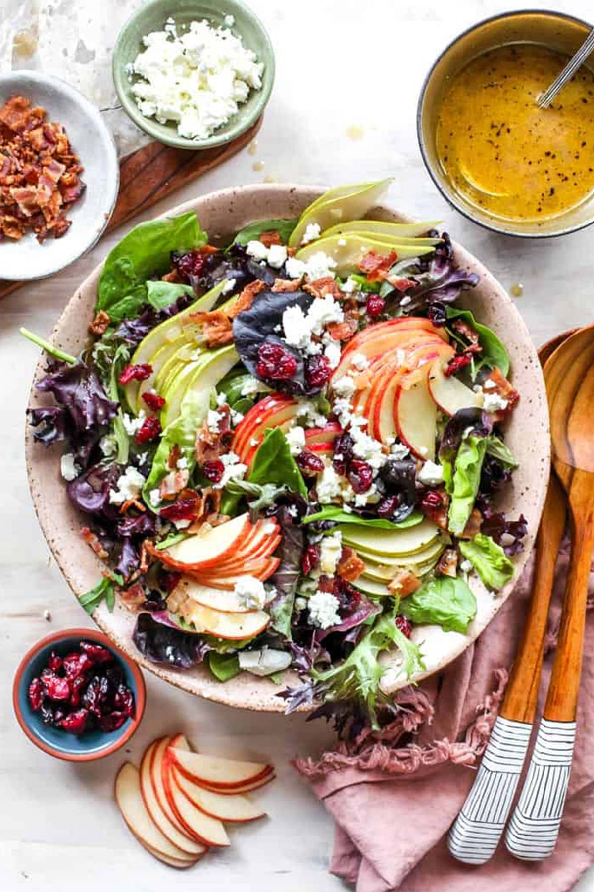 A pink bowl of autumn salad surrounded by dried cranberries, apple cider vinaigrette and bacon with spoons nearby