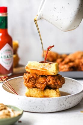 A fried chicken breast is between two cornmeal waffle pieces with maple syrup being poured over it