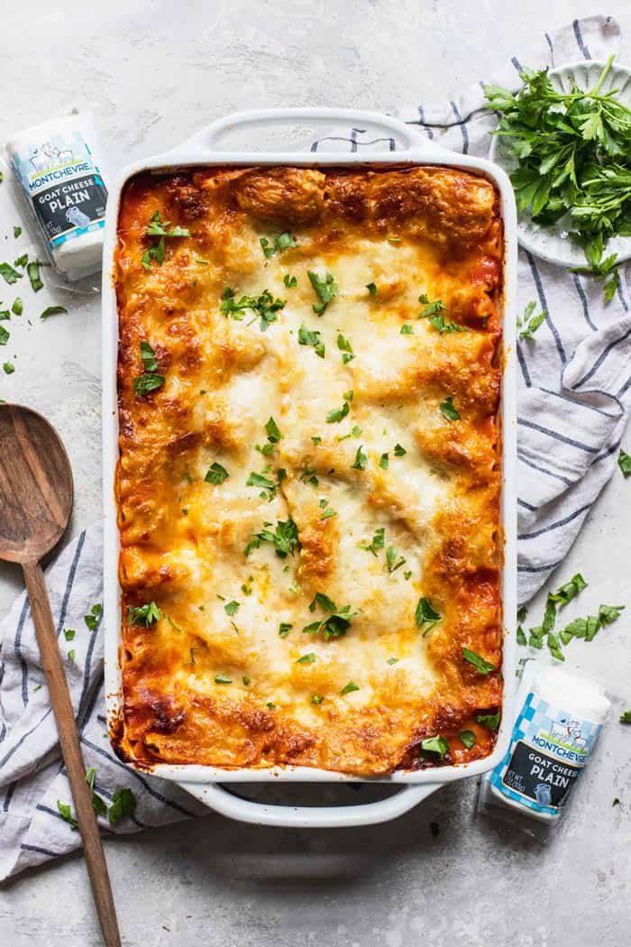 A large pan of baked lasagna sprinkled with parsley against a white background with parsley in a bowl and goat cheese on the side
