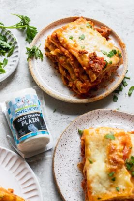 Two big slices of lasagna on white plates next to a roll of goat cheese and parsley on a plate