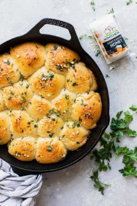 A cast iron skillet filled with cheese stuffed rolls with a garlic butter sauce on top next to goat cheese and herbs
