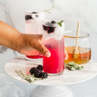 Hand picking up a refresher glass to drink next to honey, berries and sage leaves on a white platter