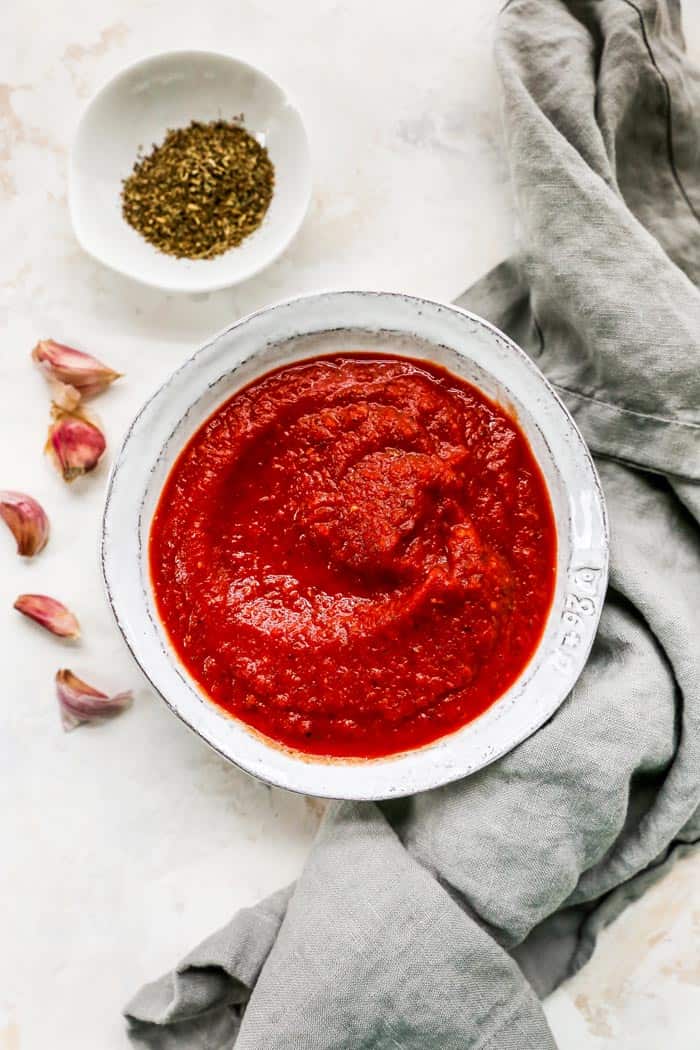 A white bowl of pizza sauce just prepared against a gray napkin with herbs and garlic