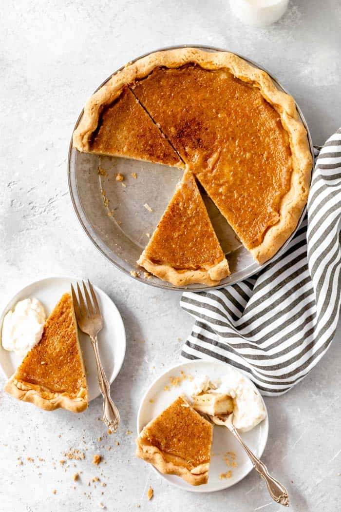 A top down image of a sliced Southern egg pie with slices on white plates with whipped cream next to a striped napkin