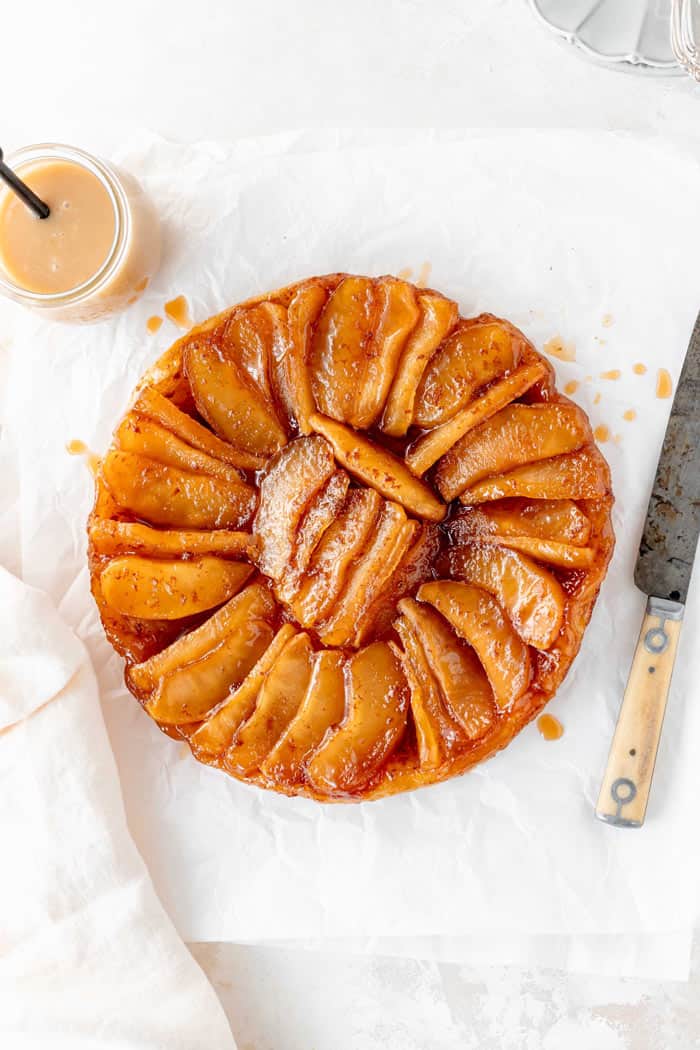 An apple tartin after just being flipped with a knife and cream sauce against white background