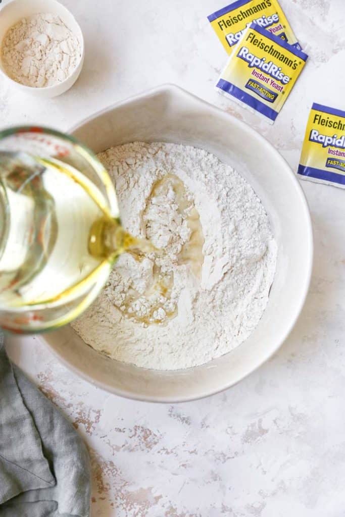oil being poured into flour and instant yeast in a large bowl to make pizza dough