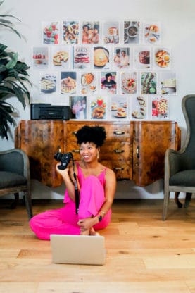 Jocelyn Delk Adams sitting on a floor holding a Canon camera n her laptop working with images of her brand on the wall behind her