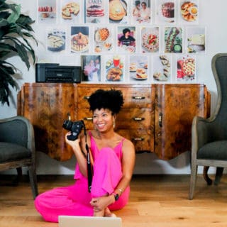 Jocelyn Delk Adams sitting on a floor holding a Canon camera n her laptop working with images of her brand on the wall behind her