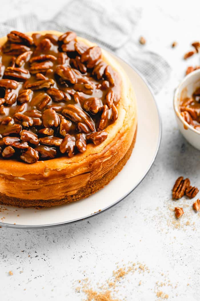 A close up of a pecan pie cheesecake on a white plate with pecans scattered on the white background