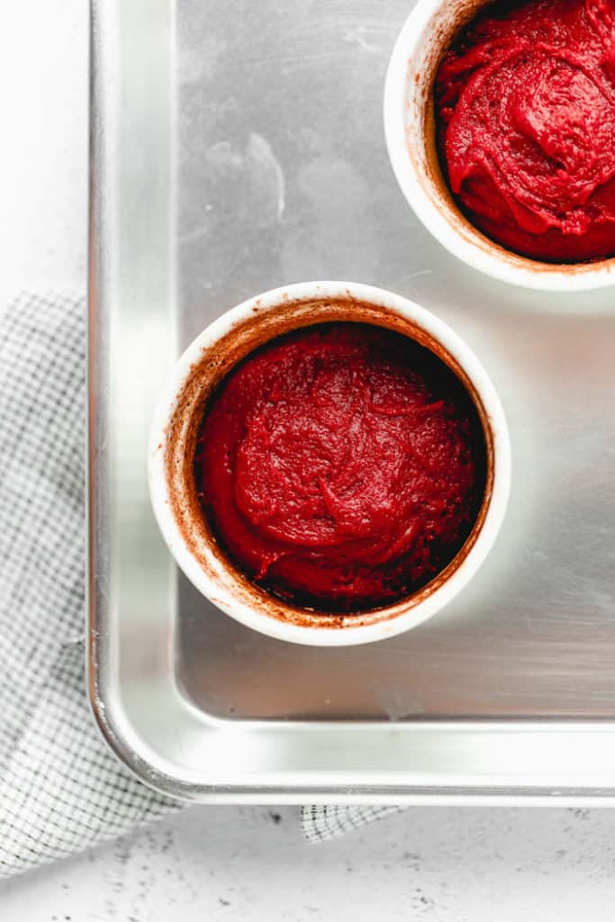 White ramekins on a baking sheet with red velvet batter inside of them ready to bake