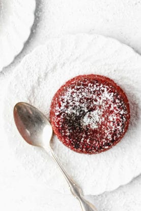 A red velvet molten cake out of the oven on a white plate topped with confectioner's sugar with a spoon nearby