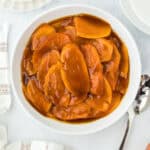 A white plate of candied sweet potatoes glazed on a white background with a spoon and sweet potatoes as well.