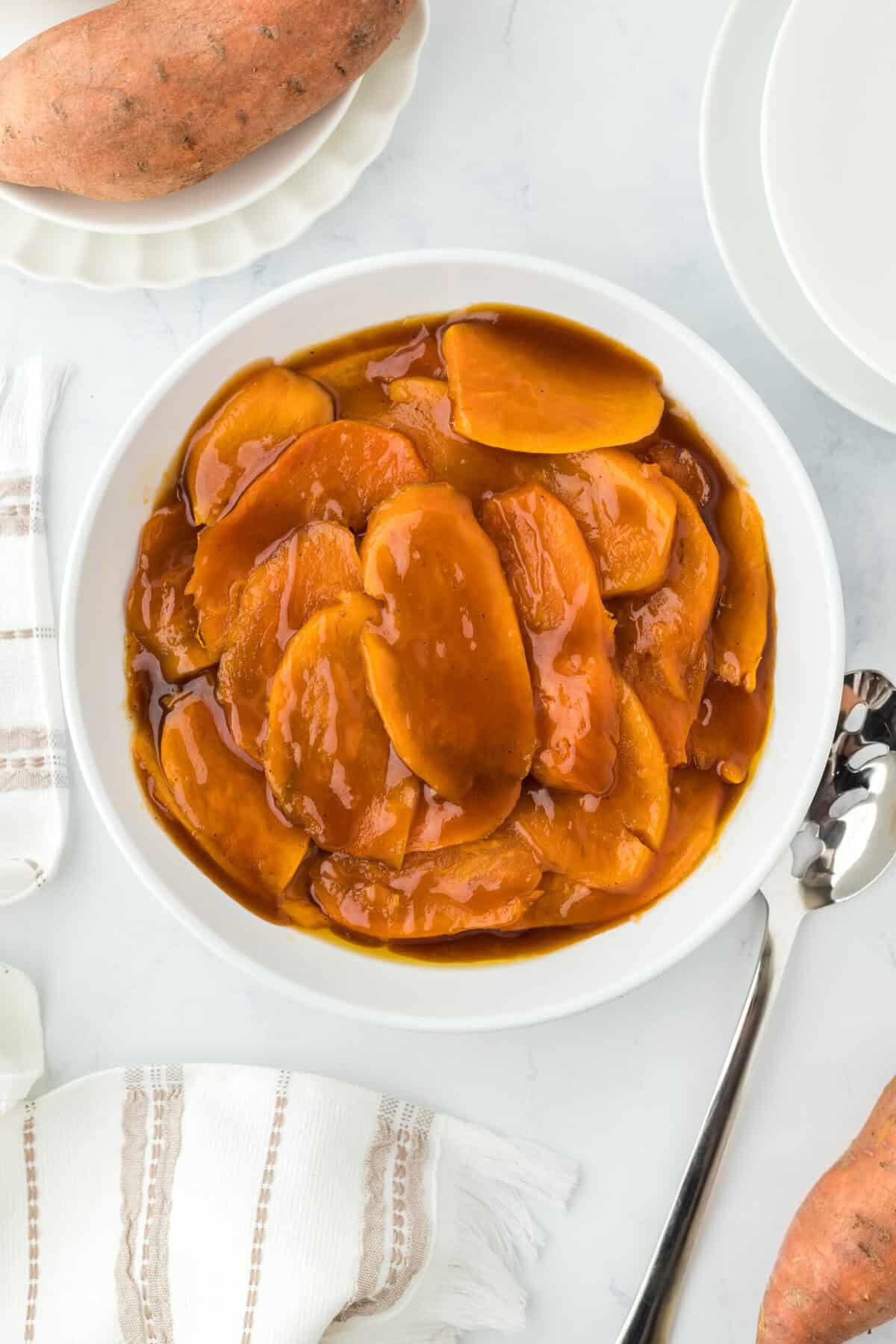 A white plate of candied sweet potatoes glazed on a white background with a spoon and sweet potatoes as well.