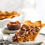 A slice of sweet potato pecan pie on a white plate with fork next to whole pie and small bowl of pecans