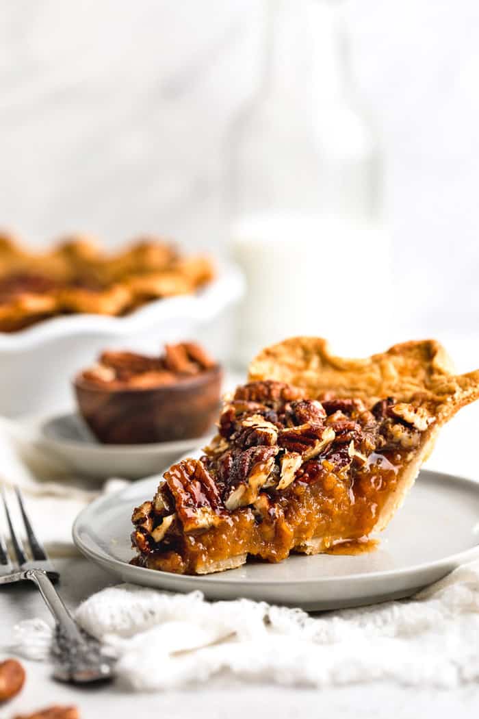 A slice of sweet potato pecan pie on a white plate with fork next to whole pie and small bowl of pecans