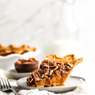 A slice of sweet potato pecan pie on a white plate with fork next to whole pie and small bowl of pecans