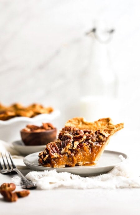 A slice of sweet potato pecan pie on a white plate with fork next to whole pie and small bowl of pecans