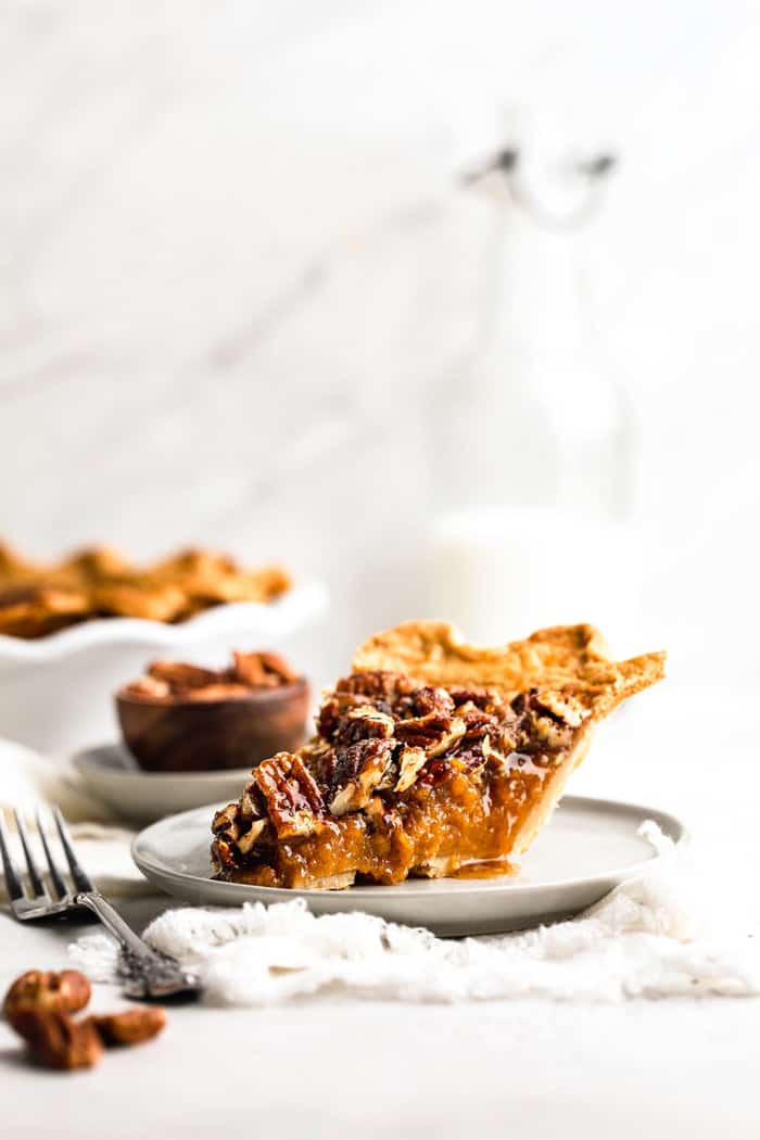 A slice of sweet potato pecan pie on a white plate with fork next to whole pie and small bowl of pecans