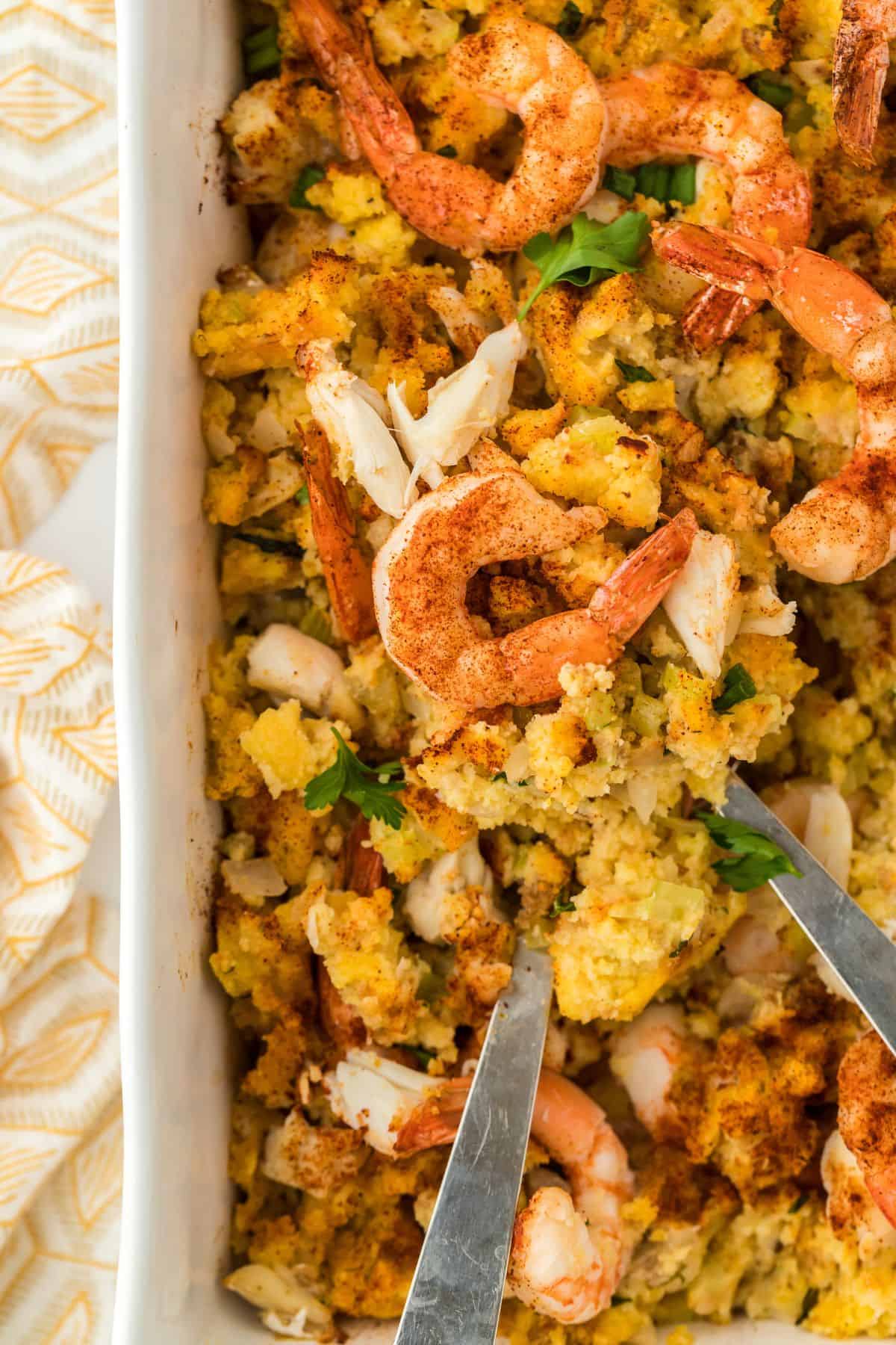 A baking dish filled with seafood dressing, topped with plump shrimp and crab meat, with two spoons inside