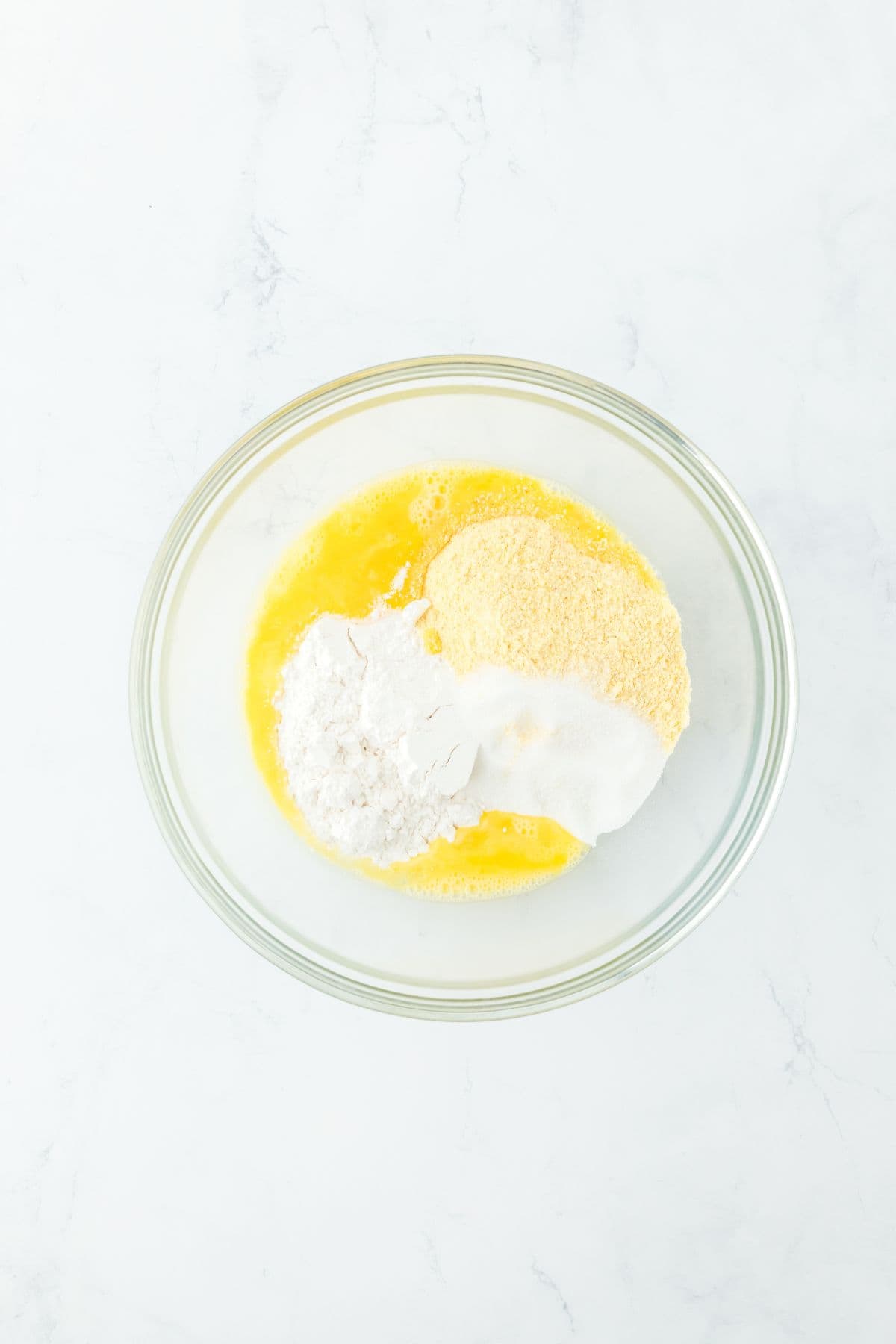 A glass bowl containing cornbread mix, flour, sugar, and beaten eggs, ready for mixing