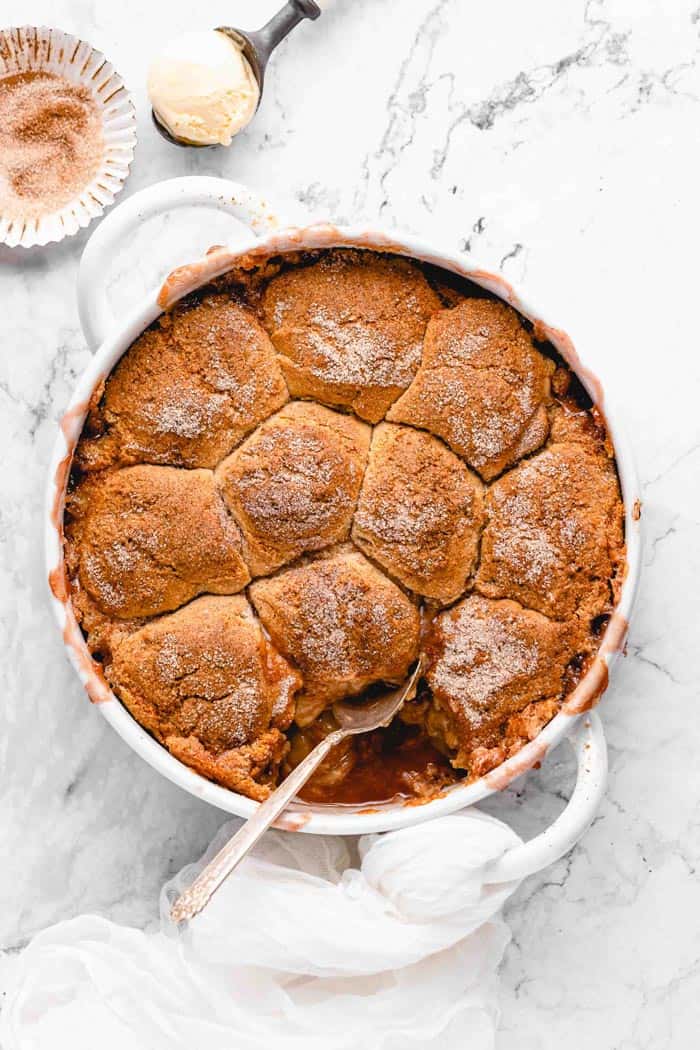 A freshly baked apple cobbler in a white pan with a spoon in the center scooping some out