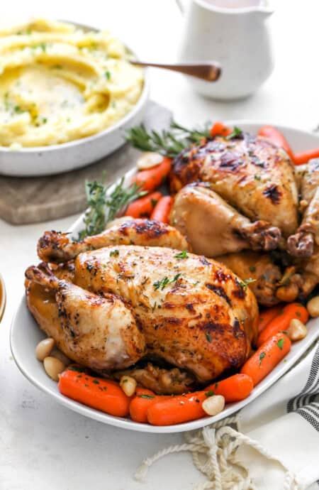 Two spiced cornish hens on a white platter with mashed potatoes and carrots
