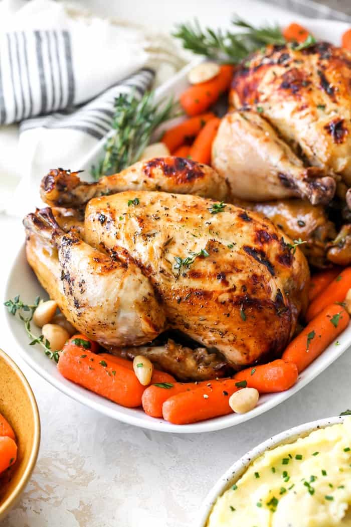 A close up of cider marinated cornish hens on a white platter with carrots and white towel in background
