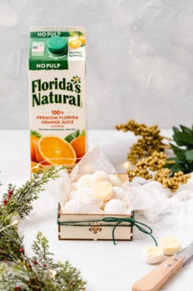 A holiday gift box filled with white chocolate orange truffles against a holiday background with a carton of orange juice