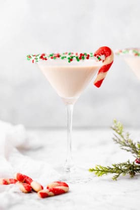 A tall glass of a sugar cookie martini with sprinkles around the rim and a candy cane shaped cookie ready to serve for the holidays