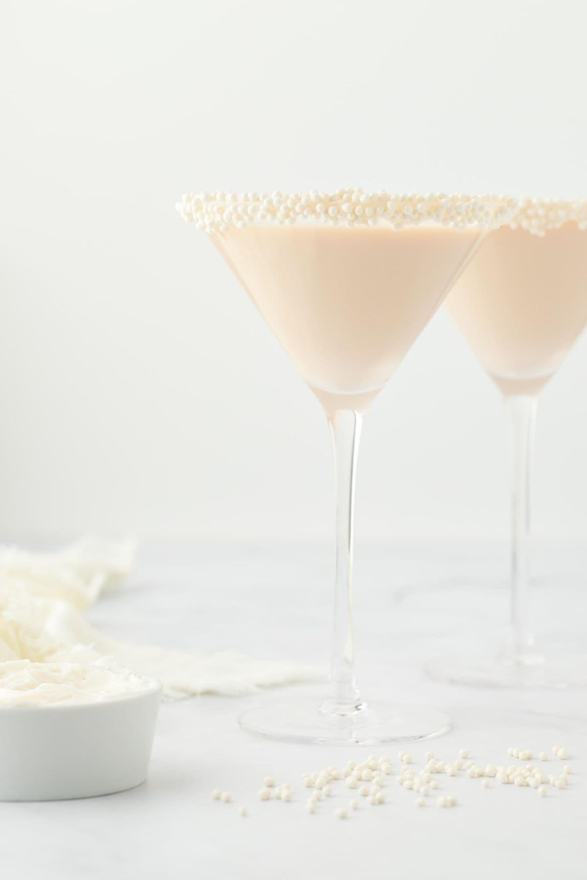 Two martini glasses with sugar pearl-sprinkled rims filled with sugar cookie martini, alongside a small bowl of white frosting and scattered sugar pearls, set against a white background