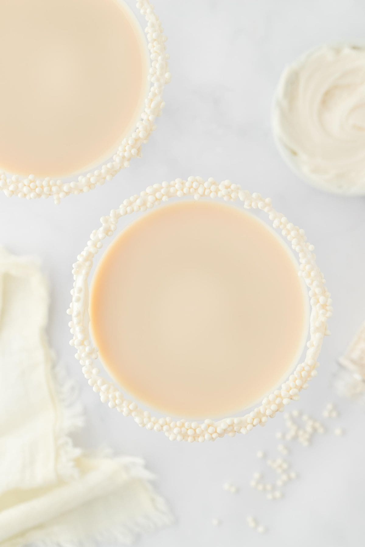 Overhead shot of a martini glass with sugar cookie martini and with a pearl-sprinkled rim, placed next to a small bowl of white frosting and scattered sugar pearls