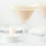 Two martini glasses with sugar pearl-sprinkled rims filled with sugar cookie martini, alongside a small bowl of white frosting and scattered sugar pearls, set against a white background