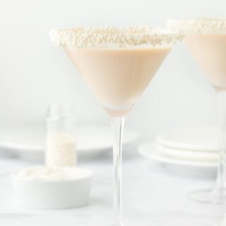 Two martini glasses with sugar pearl-sprinkled rims filled with sugar cookie martini, alongside a small bowl of white frosting and scattered sugar pearls, set against a white background