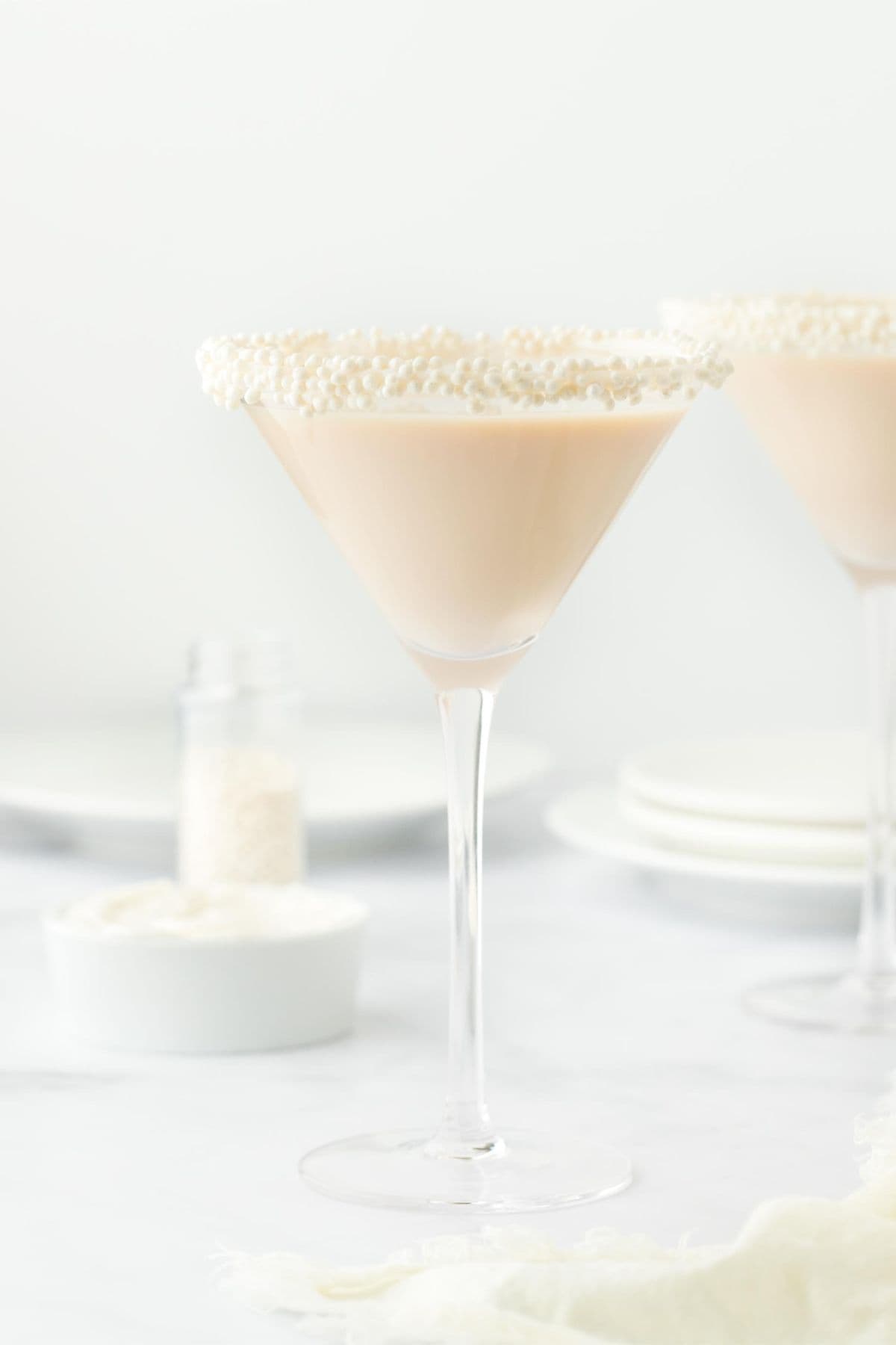 Two martini glasses with sugar pearl-sprinkled rims filled with sugar cookie martini, alongside a small bowl of white frosting and scattered sugar pearls, set against a white background