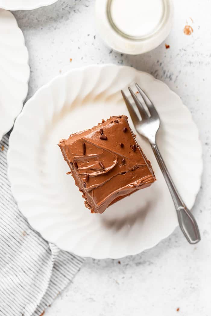 A slice of sheet cake with chocolate frosting on a white plate against a white background