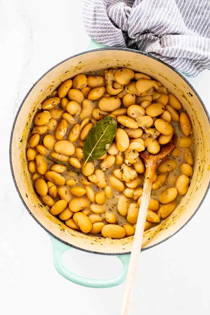 A wooden spoon in a large white pot of butter beans with a bay leaf on top