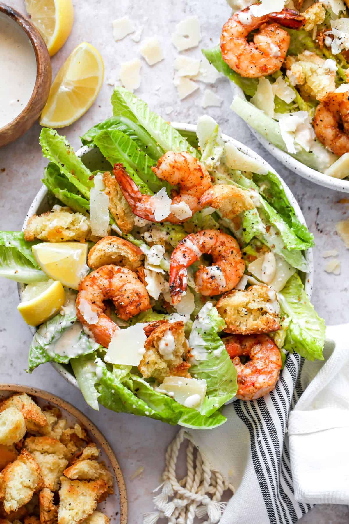 A bowl of Caesar salad topped with grilled shrimp, croutons, shaved parmesan, and Caesar dressing, with lemon wedges on the side