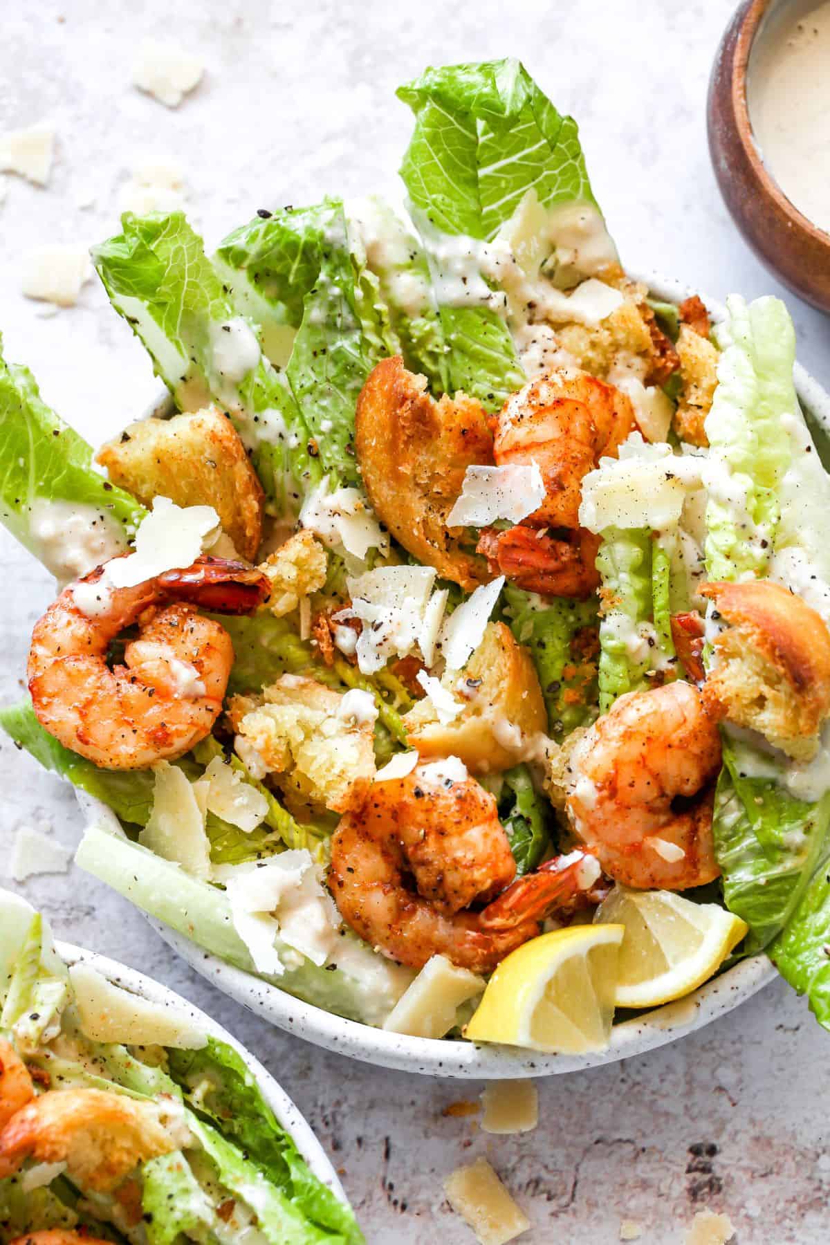 Bowl of shrimp Caesar salad generously drizzled with creamy dressing and garnished with shaved parmesan and black pepper