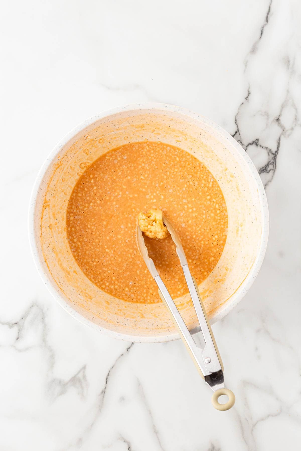 Cauliflower batter in a bowl with tongs holding one coated floret