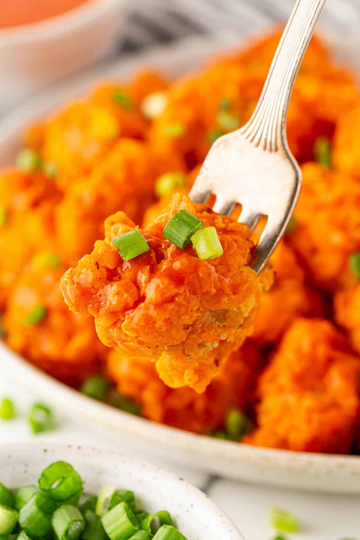 A fork holding a saucy buffalo fried cauliflower bite topped with sliced green onions