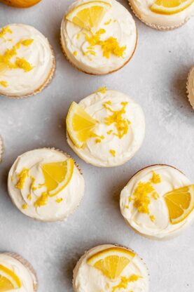 An overhead photo of several perfectly frosted lemon cupcakes with lemon wedges and zest on top