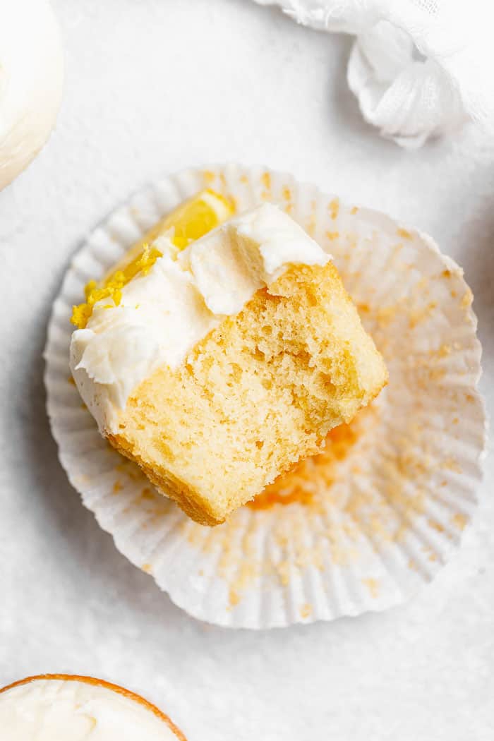 A close up of lemon cupcake with big bite taken against open wrapper on white background