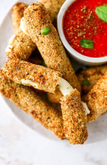 A delicious close up of mozzarella sticks on a white plate with one showing cheese pull