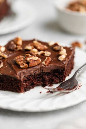 A close up of chocolate sheet cake recipe on white plate with moist texture of cake inside with some eaten with nearby fork
