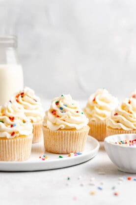 A white plate filled with cupcakes with a vanilla buttercream and sprinkles