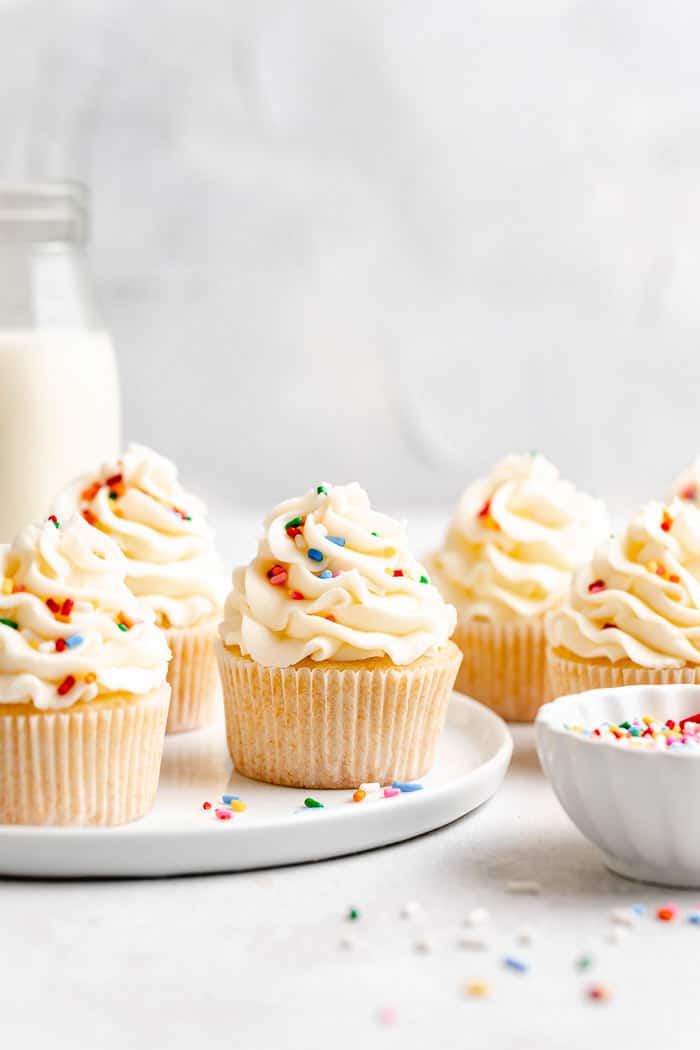 A white plate filled with vanilla cupcakes with a vanilla buttercream and sprinkles on a white background