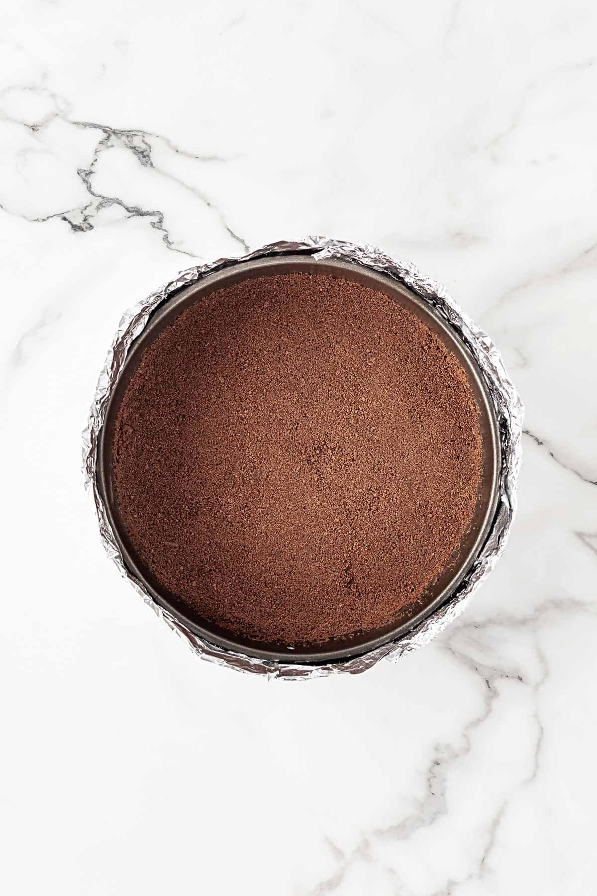 Chocolate cookie crust pressed evenly into a foil-wrapped springform pan on a marble surface
