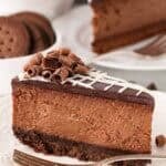 A slice of chocolate cheesecake with chocolate cookie crust topped with chocolate curls on a white plate with a fork next to it and cheesecake slices, chocolate, and cookies in the background
